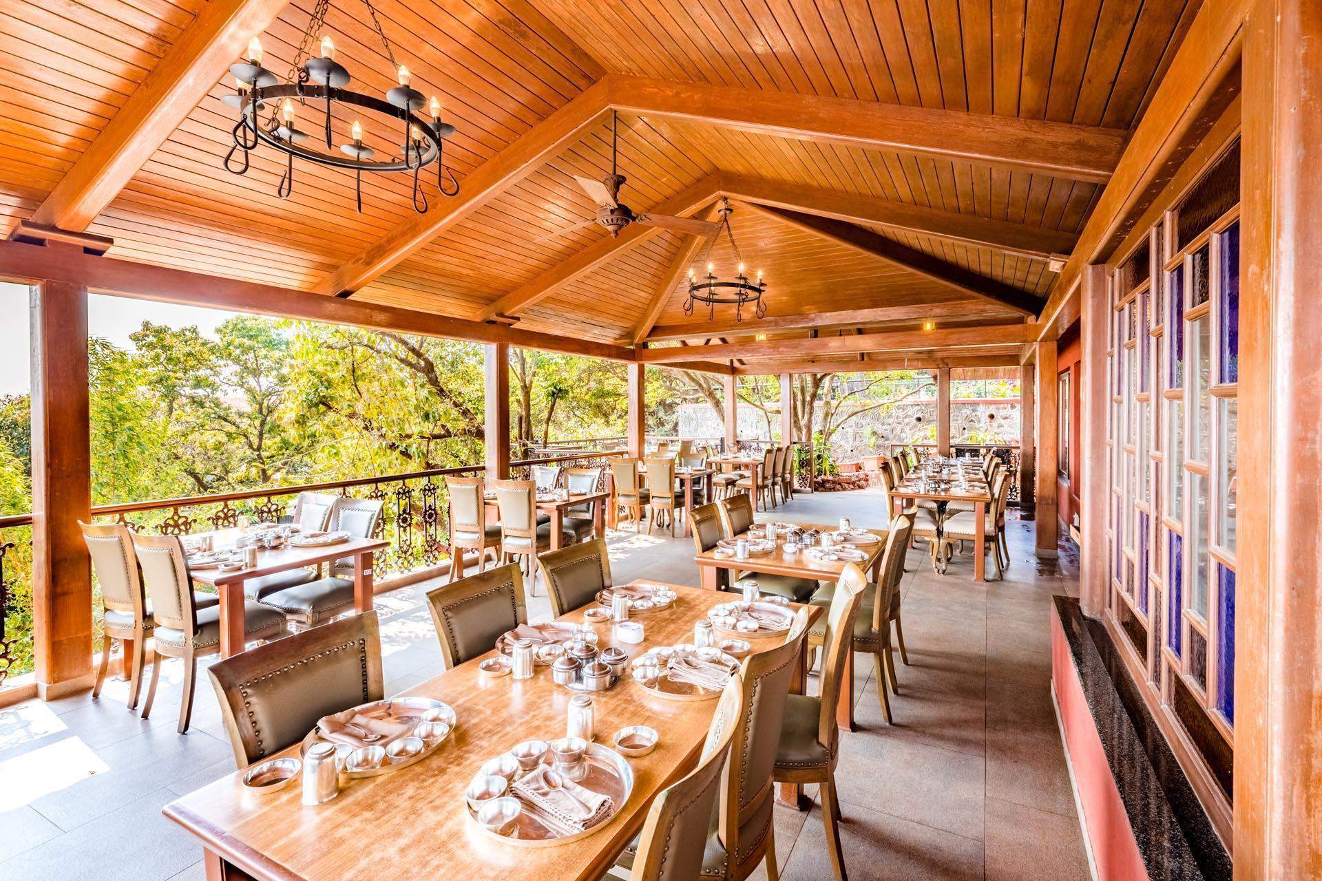 An open-air restaurant at Ramsukh Resort with wooden furniture, a high ceiling, and views of the surrounding greenery, offering a cozy dining experience.